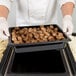A person in white gloves holding a Cambro black plastic food pan full of meatballs.