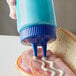 A hand using a Vollrath blue Twin Tip squeeze bottle to pour mayonnaise onto a sandwich with ham.