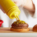 A person pours yellow mustard from a squeeze bottle onto a burger.