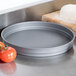 An American Metalcraft hard coat anodized aluminum cake pan on a counter next to pizza dough and tomatoes.