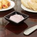 A table with a bowl of pink yogurt and a Carlisle black square ramekin with a silver spoon.
