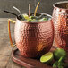 Two copper mugs with ice, limes, and metal bent straws on a wooden surface.