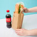 A hand holding a Duro brown paper bag with a sandwich and a soda bottle inside.