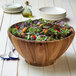 An acacia wood salad bowl filled with salad on a table with utensils.