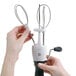 A hand using an OXO stainless steel manual crank egg beater with a black rubber handle.