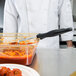 A person in a white coat using a Vollrath black perforated oval Spoodle to serve red sauce from a pan.