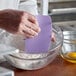 A person using a Choice purple plastic bowl scraper to mix batter in a glass bowl.