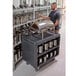 A man standing by a Cambro charcoal gray service cart full of silver containers.