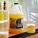 A jug of Finest Call sweet and sour concentrate on a yellow table next to a glass of yellow liquid.