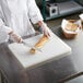 A chef in a white coat uses a Thunder Group white polyethylene cutting board to cut a loaf of bread.