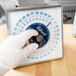 A person in a white glove uses an Edlund SR-2 mechanical portion scale on a counter.