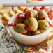 A white bowl of Belosa Turkish Red Pepper Stuffed Queen Olives on a table with cheese and grapes.