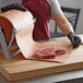 A person in a red apron using 12'' PeachTREAT Butcher Paper to wrap meat on a table.