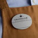 A brown apron with a customizable oval silver plastic nametag with the Coffeeco logo.