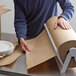 A person rolling a Lavex natural kraft void fill packing paper roll on a table.