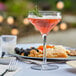 A Libbey Tritan plastic coupe glass filled with pink liquid and garnished with rosemary on a table with a plate of food.