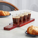 An Acopa mahogany flight paddle holding three shot glasses of brown liquid on a wooden board with a croissant and a plate of food.