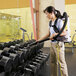 A woman using a ProTeam backpack vacuum to clean dumbbells in a gym.