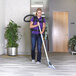 A woman in a purple shirt and purple backpack using a ProTeam straight telescoping wand to vacuum a room.