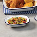An American Metalcraft white rectangular bowl with blue rim filled with fried chicken, corn, and coleslaw.