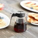A Tablecraft glass syrup dispenser with a black lid on a table with pancakes and a drink.