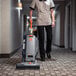 A woman in uniform using a Hoover commercial bagged upright vacuum cleaner to clean a hallway.