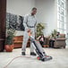 A man using a Hoover commercial upright vacuum cleaner in a room with a potted plant.