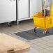 A yellow mop bucket with handle in a kitchen sitting on a white rectangular floor trough grate.