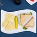 A white Fineline plastic luncheon plate with a sandwich, potato chips, and a pickle.