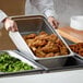 A person holding a Choice stainless steel steam table pan of fried chicken in front of a hotel buffet.