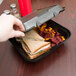 A hand taking out a sandwich from a Genpak black foam container.
