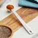 A Vollrath stainless steel round measuring spoon filled with red powder on a wooden surface.