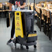 A person standing behind a Rubbermaid cleaning cart with a yellow sign on it.