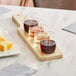 An Acopa natural wood paddle with stemless wine tasting glasses on a brown surface with cheese.