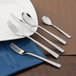 A brown surface with a blue plate with a Walco Audition stainless steel butter knife and fork.