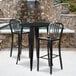 A black metal Flash Furniture bar table with two black metal chairs with vertical slat backs.