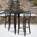 A Flash Furniture black metal bar table and two chairs on an outdoor patio in front of a stone wall.