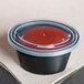 A Newspring black oval plastic souffle container with a clear lid containing red liquid.