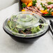 A bowl of salad in a black plastic bowl with a clear plastic lid.