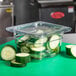 A clear Cambro lid on a glass container of cut green cucumbers.