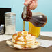 A person using a Tablecraft teardrop syrup dispenser to pour syrup on a stack of pancakes.