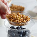 A hand holding a bowl of granola and blueberries over a clear plastic parfait insert.