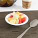 A Carlisle white melamine bowl filled with fruit with a spoon next to it.