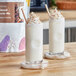 A white cylinder filled with Bossen Almond Powder Mix next to two glasses of milkshakes.