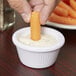 A person's fingers holding a carrot stick and dipping it into a white Carlisle ramekin.