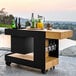 An Astella black outdoor mobile bar with bottles and glasses on it.