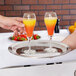 A hand holding a Vollrath Esquire stainless steel tray with two glasses of orange juice on it.