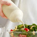 A person pouring white dressing from a clear plastic squeeze bottle onto a salad.