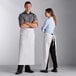 A man and woman wearing Choice white bistro aprons standing in a professional kitchen.