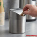 A hand putting a packet into a Vollrath stainless steel mini waste can on a counter.